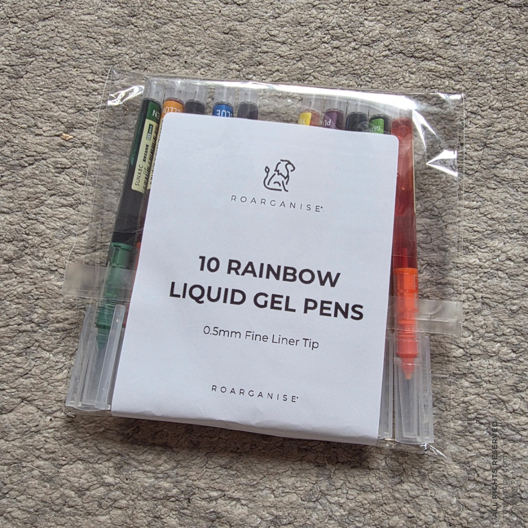A clear plastic bag containing a set of 10 liquid gel pens with a white paper insert that reads '10 RAINBOW LIQUID GEL PENS' and shows the ROARGANISE brand logo, placed on a gray carpeted floor.