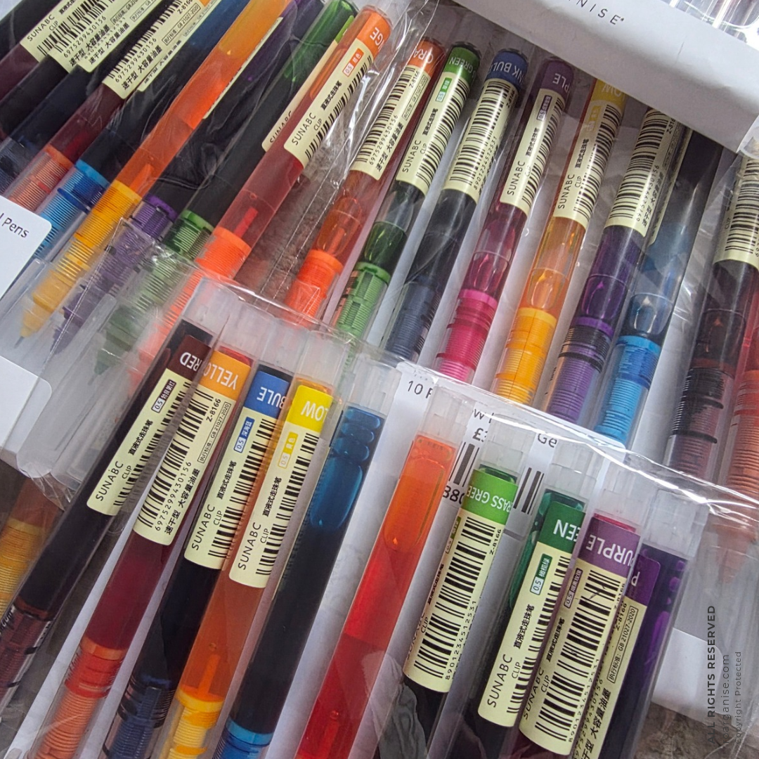 A close-up of a boxed set of multiple retractable gel pens in clear plastic packaging, showing many different ink colors and labels.